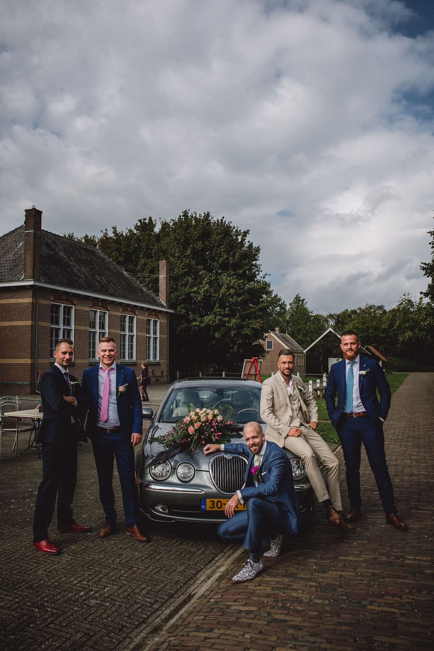 Groomsmen in navy suits pose with a vintage Jaguar decorated with flowers outside a rustic venue.Door Kim van de Broek Fotografie