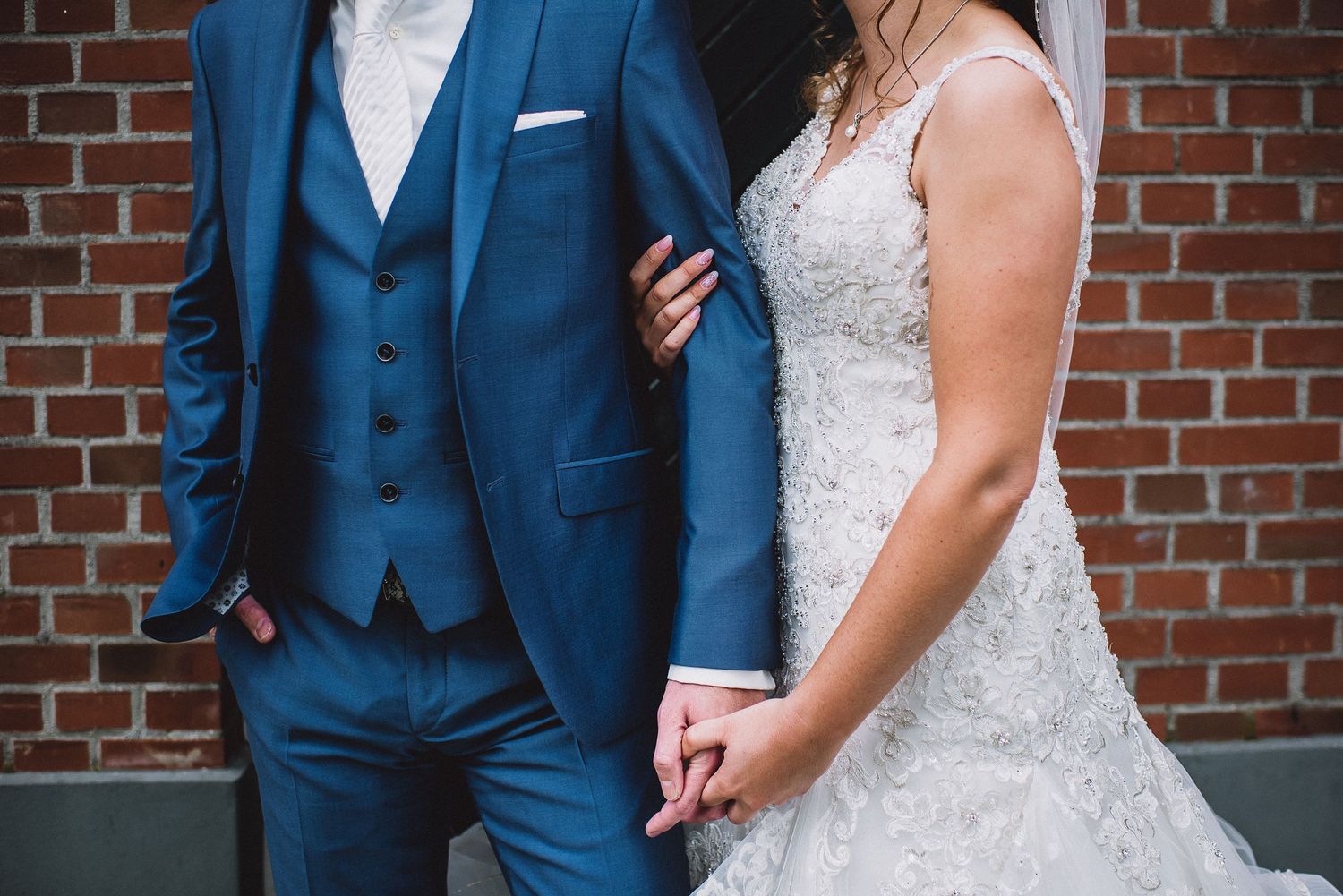 Close-up of bride in lace gown and groom in blue three-piece suit holding hands at their wedding.