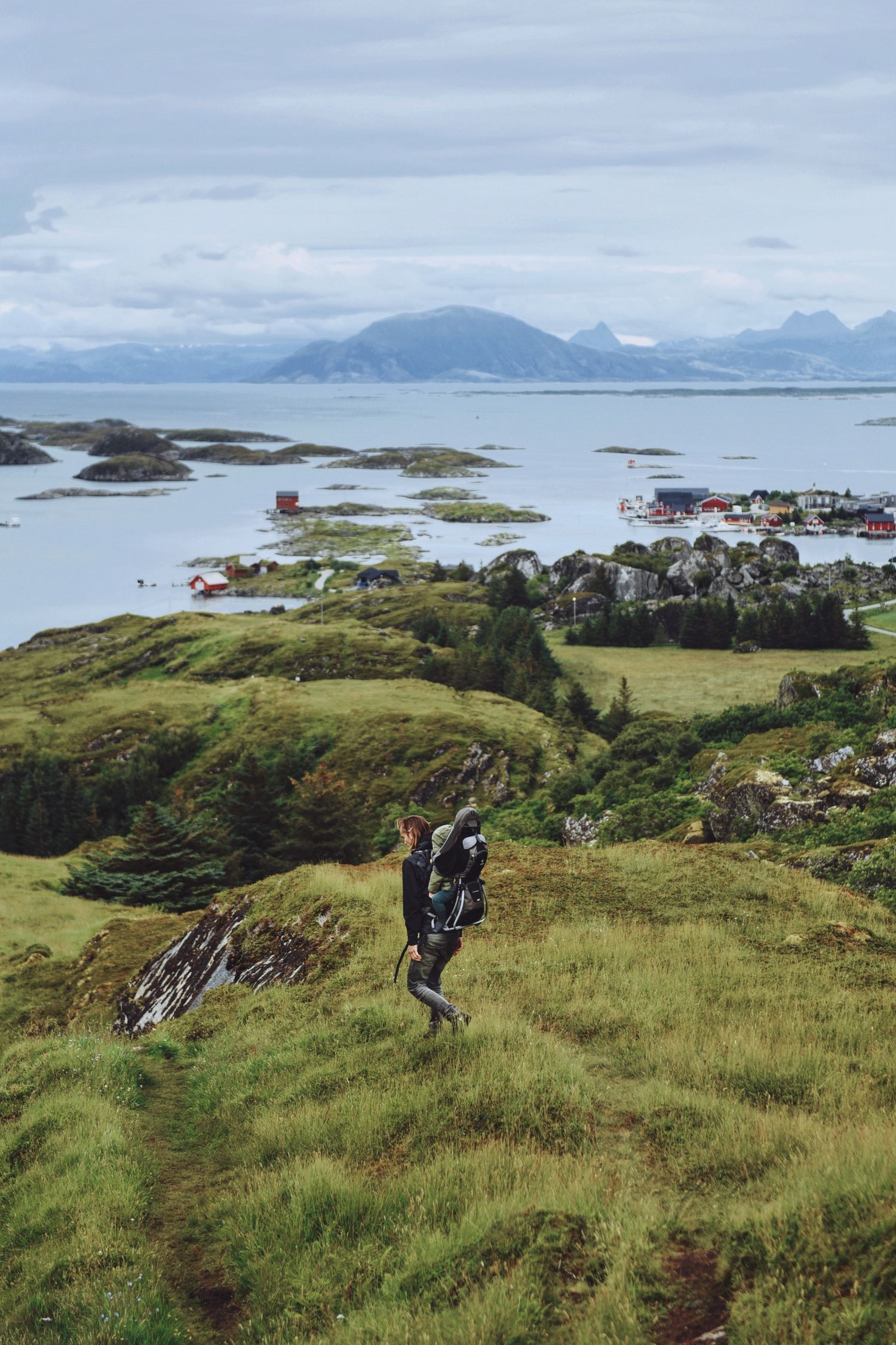 A hiker climbs a green hillside overlooking a scenic Norwegian coastal village and calm fjord waters.
