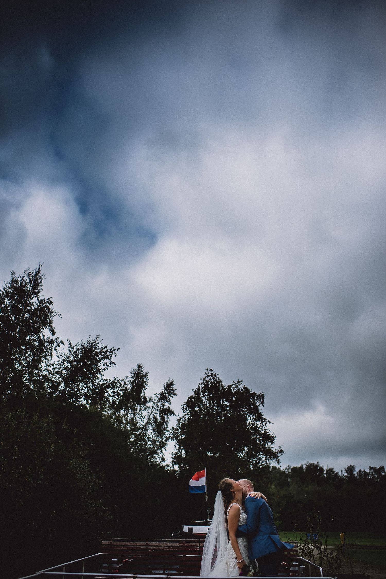 Newlyweds kissing on a dock under dramatic cloudy skies with a Dutch flag in the background.