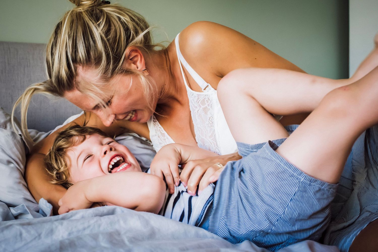 Mother and young child laughing together playfully on a bed in a bright bedroom.