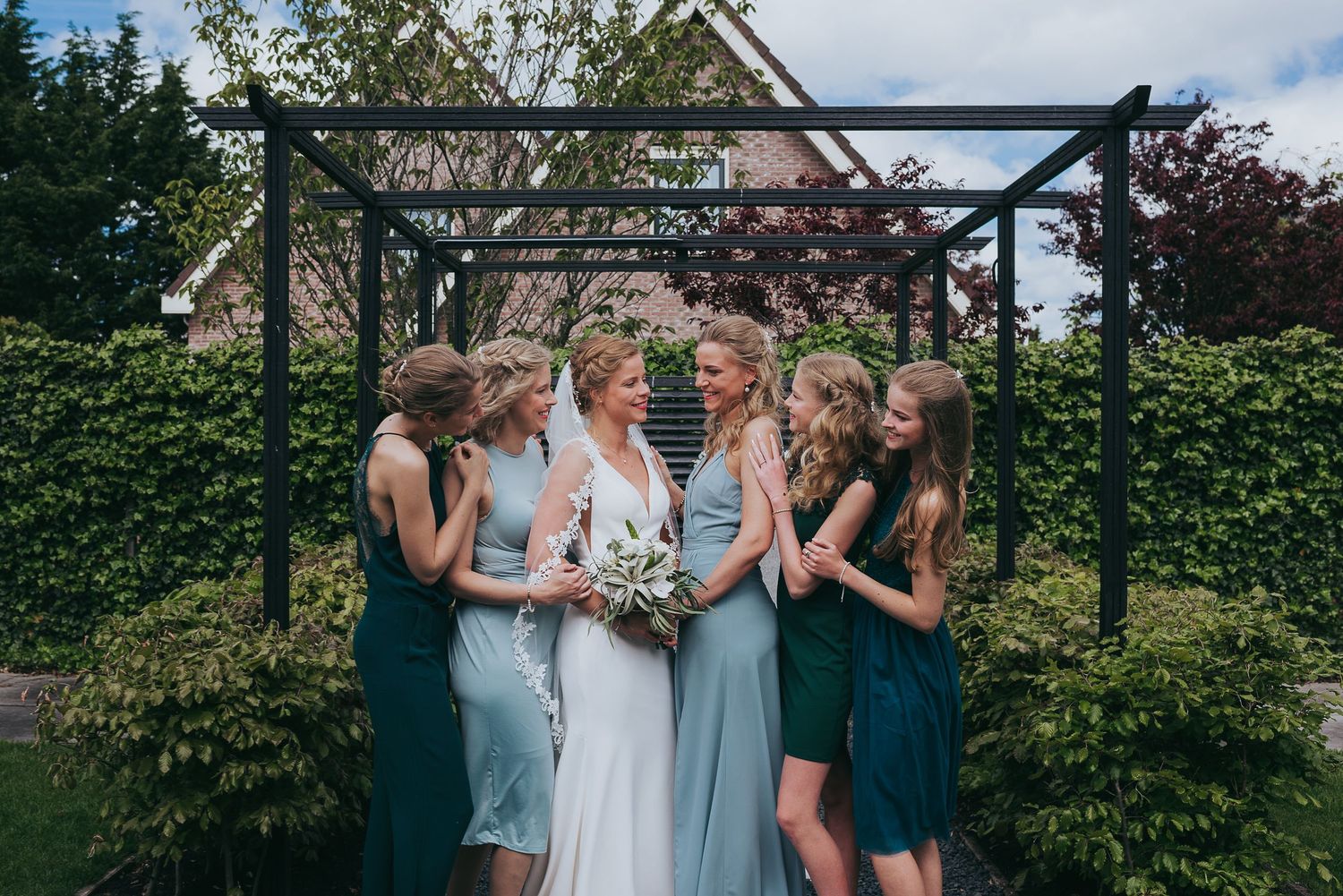 Bride laughs joyfully with bridesmaids in blue dresses under garden pergola holding bouquet.