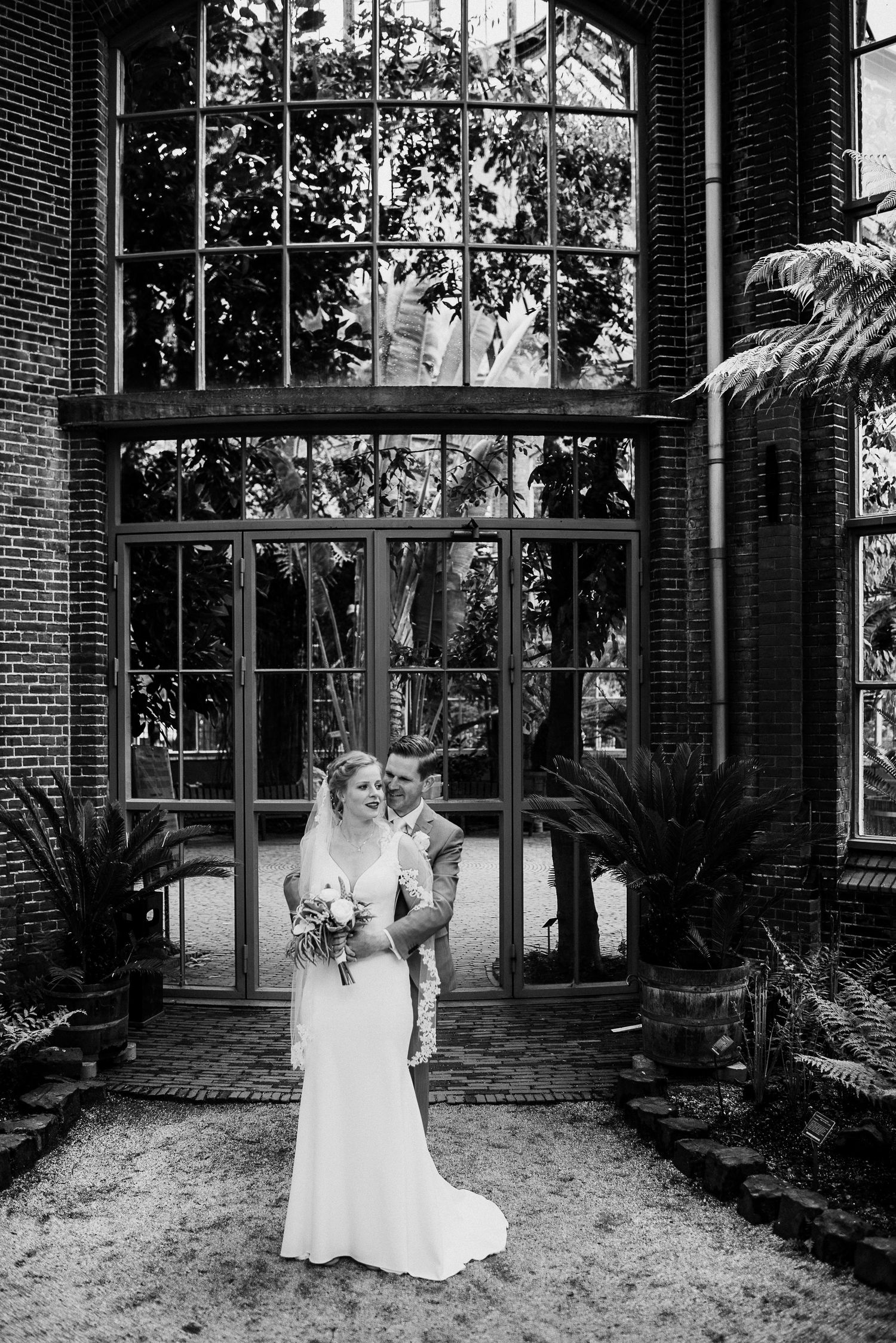 Bride and groom embracing before grand greenhouse windows surrounded by tropical plants in black and white.
