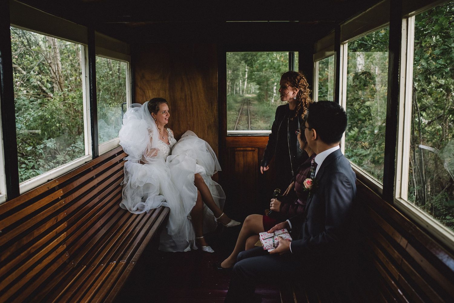 Bride in white gown sits with guests inside a wooden vintage train car surrounded by lush greenery.