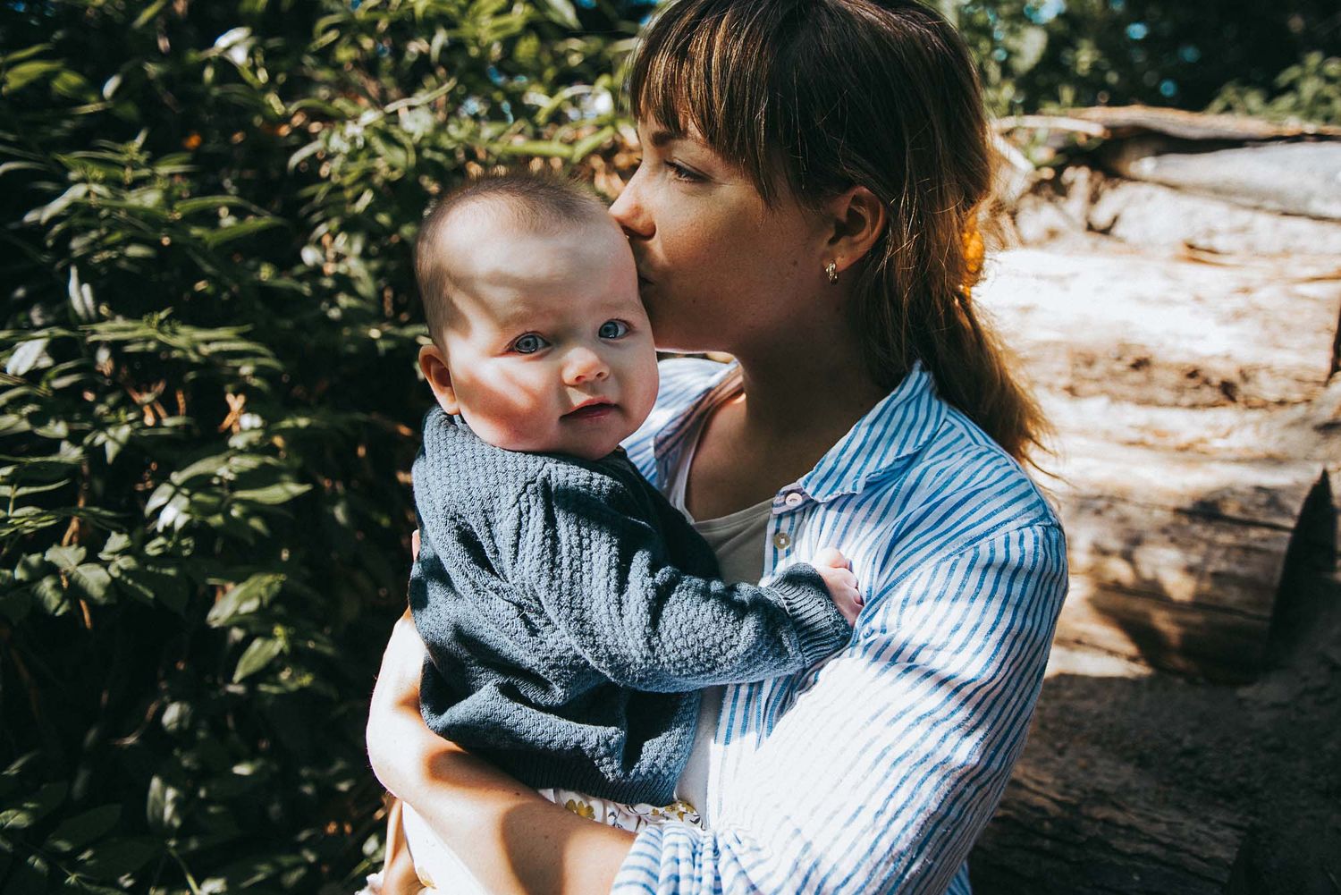 Mother kissing her newborn baby outdoors near a wooden log surrounded by green foliage.