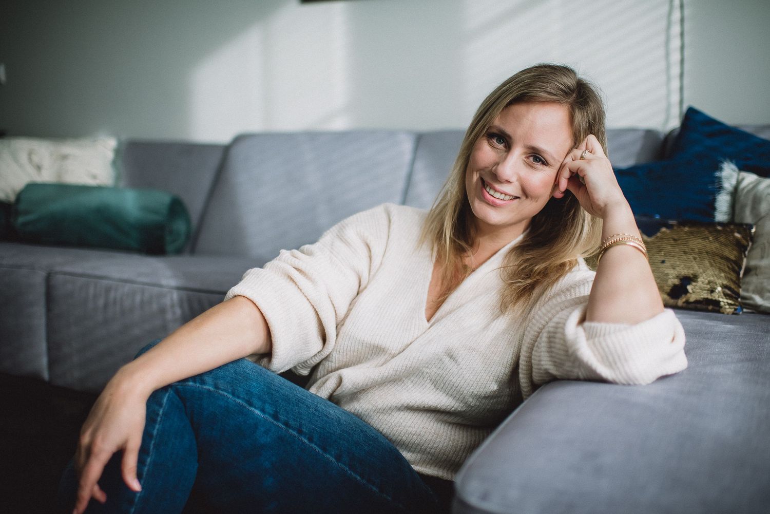 Happy blonde woman in white sweater sitting relaxed on a gray couch at home.