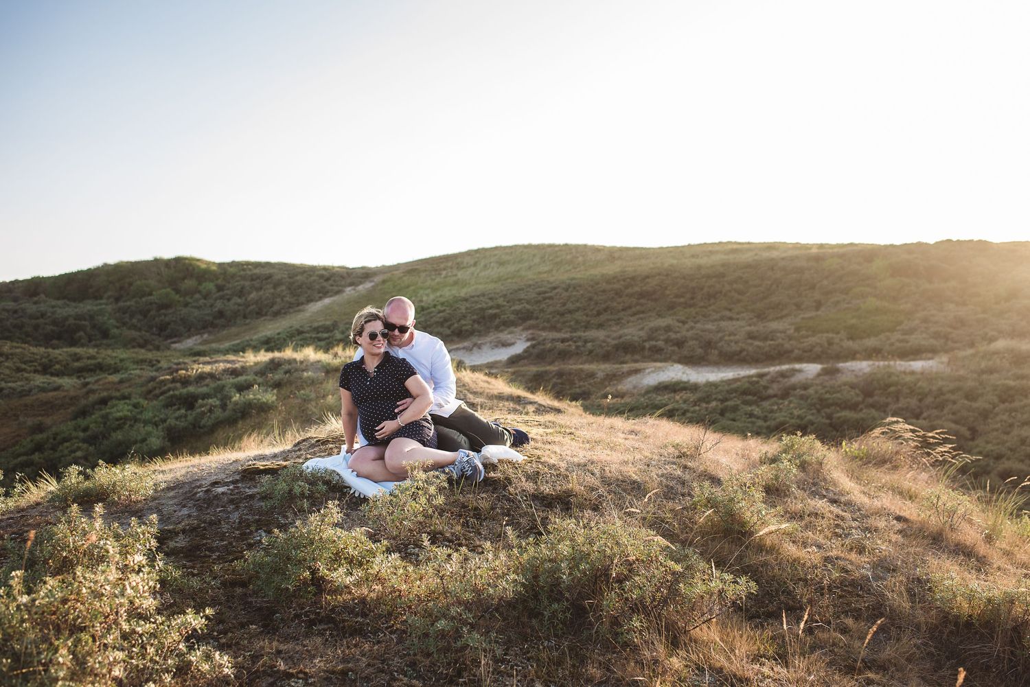 Couple sitting together on a sunlit grassy hillside enjoying golden hour.