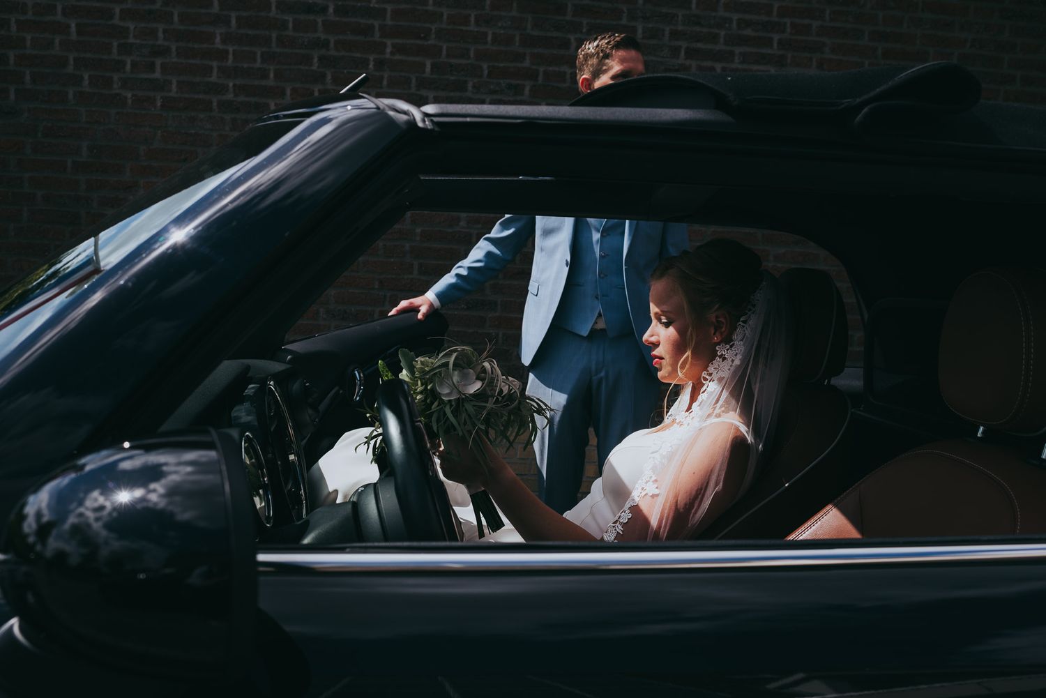 Bride sitting in convertible car holding bouquet while groom stands outside in blue suit.