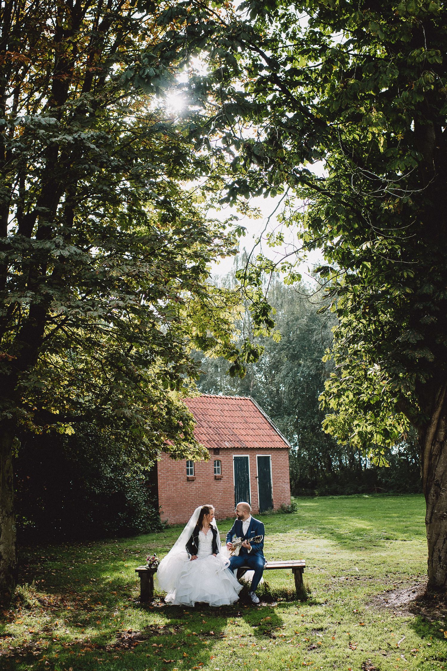 Bride and groom sit on bench beneath sunlit trees beside a charming red brick cottage in garden setting.