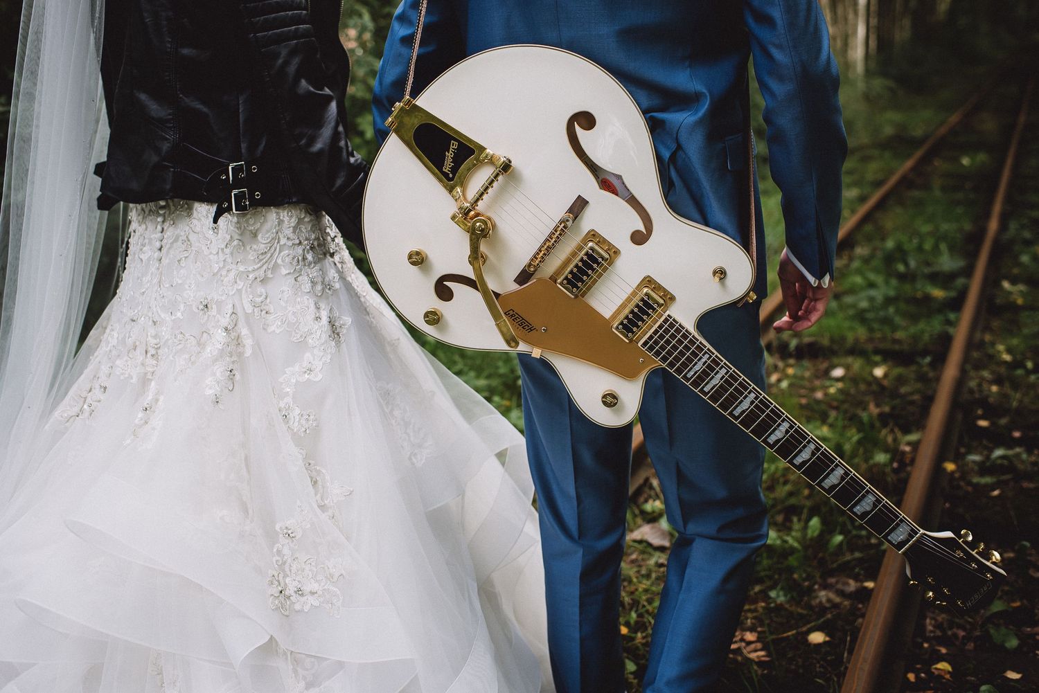 Close-up of white Gretsch guitar with gold hardware held by groom in blue suit beside bride's lace wedding dress.