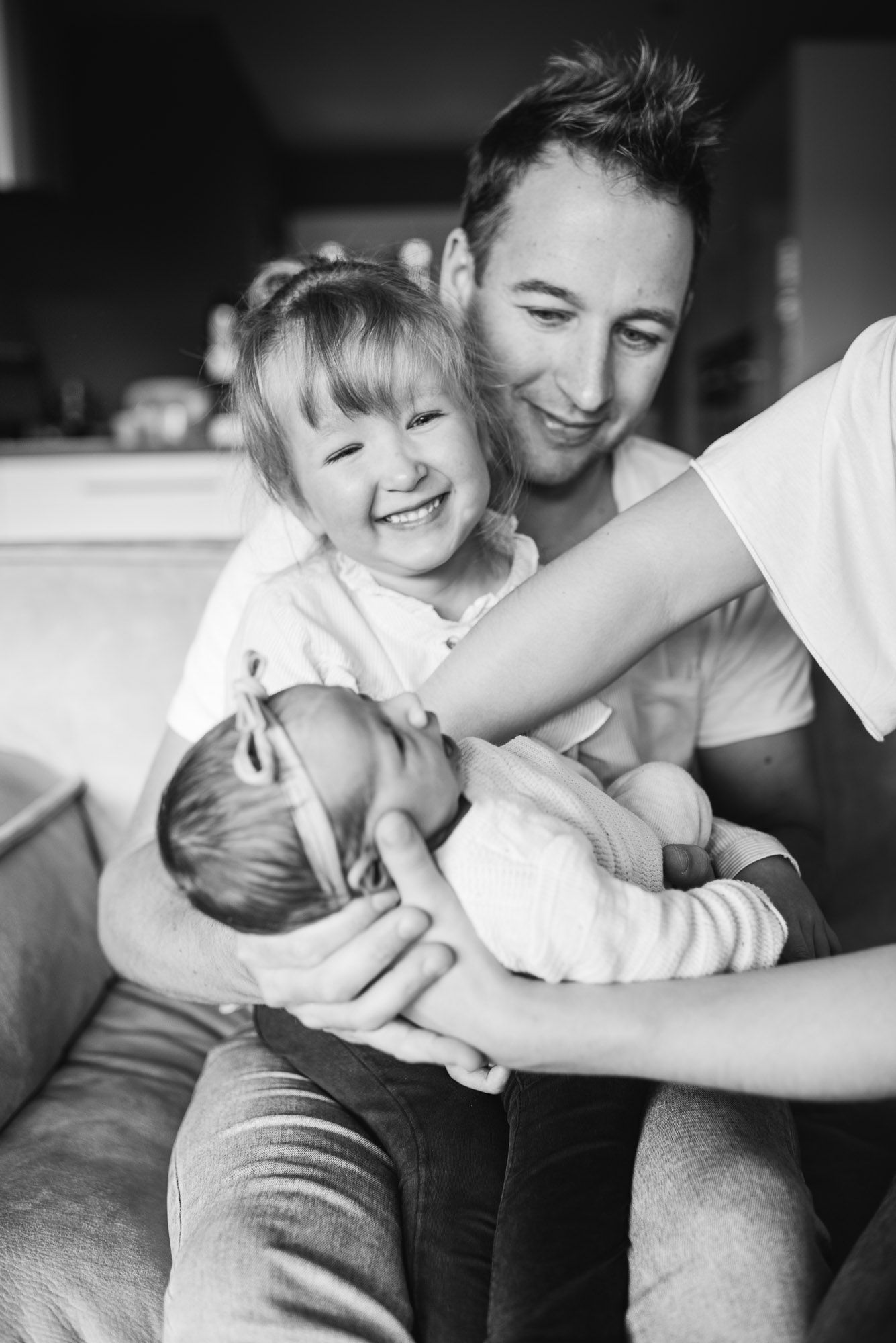 Black and white photo of a smiling father holding a newborn with an excited toddler beside him.