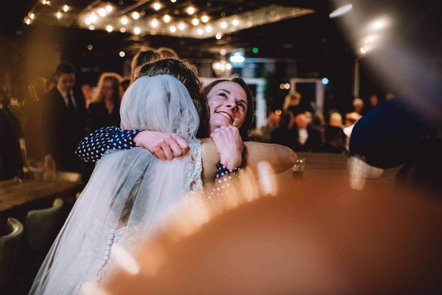 Smiling bride in veil hugging a guest at a warmly lit wedding reception. gefotografeerd door documentaire trouwfotograaf Kim van den Broek