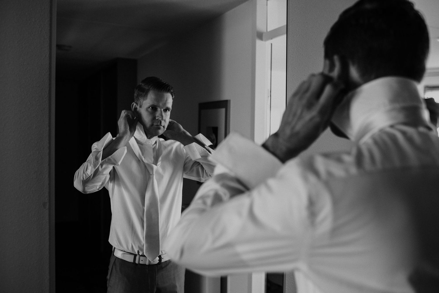 A groom adjusts his white shirt collar while looking at his reflection in a mirror before his wedding. wedding photography