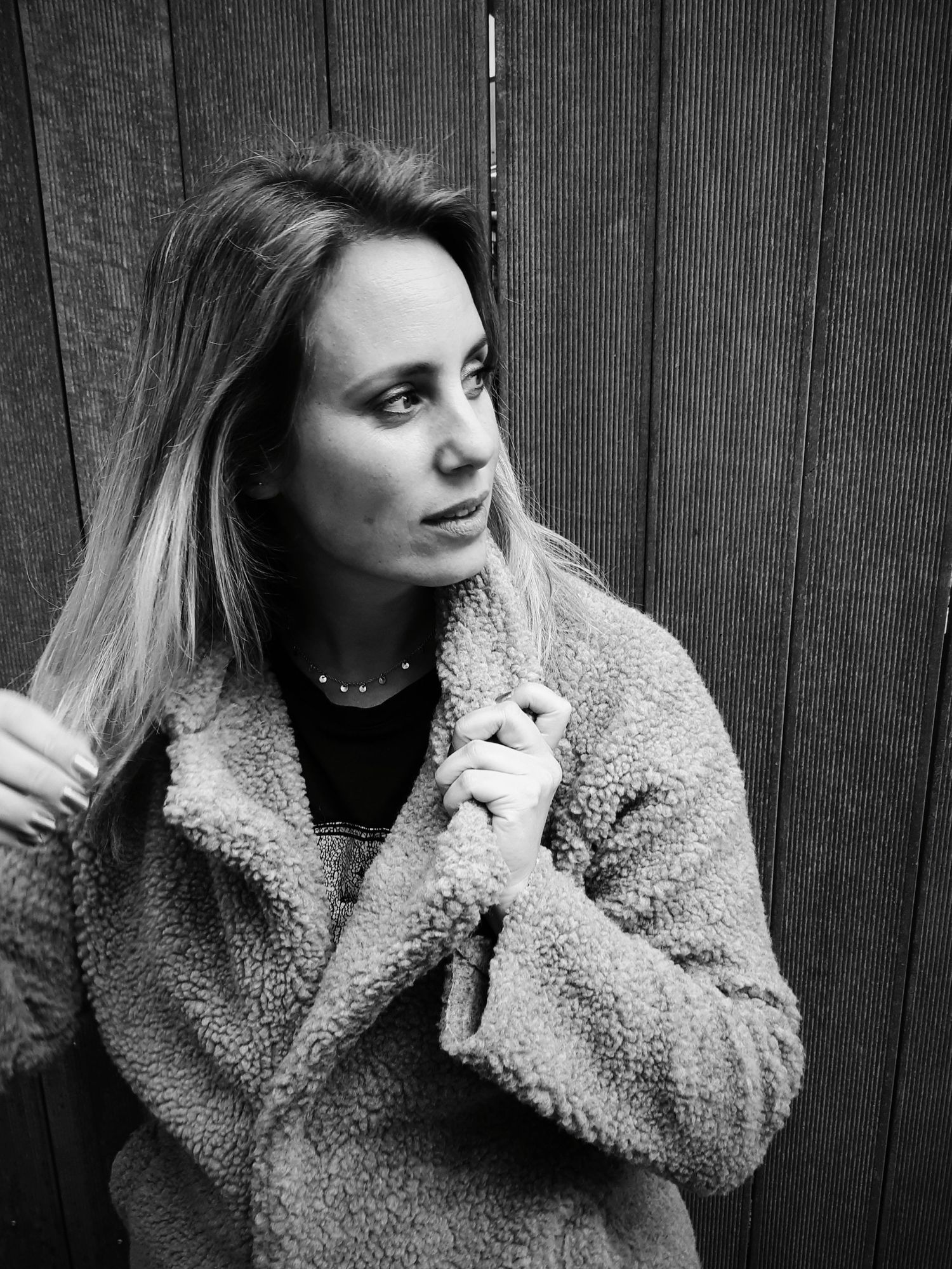 A young woman in a fluffy coat stands thoughtfully against a wooden fence in a black and white portrait. wedding photographer