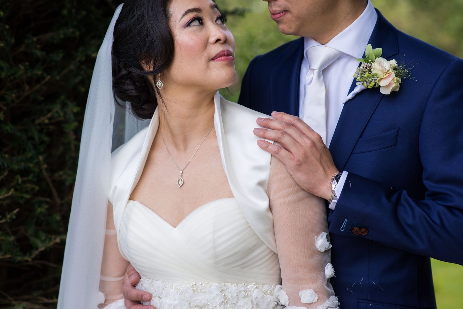 Asian bride and groom share a tender moment in wedding attire, with groom in navy suit and bride in strapless white gown.