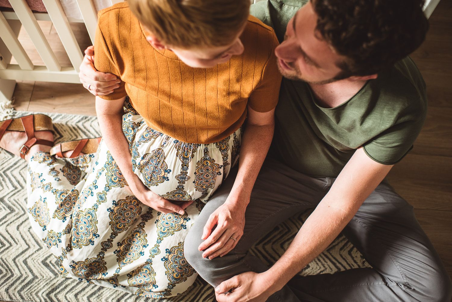 Expectant couple sitting on floor tenderly touching pregnant belly indoors.