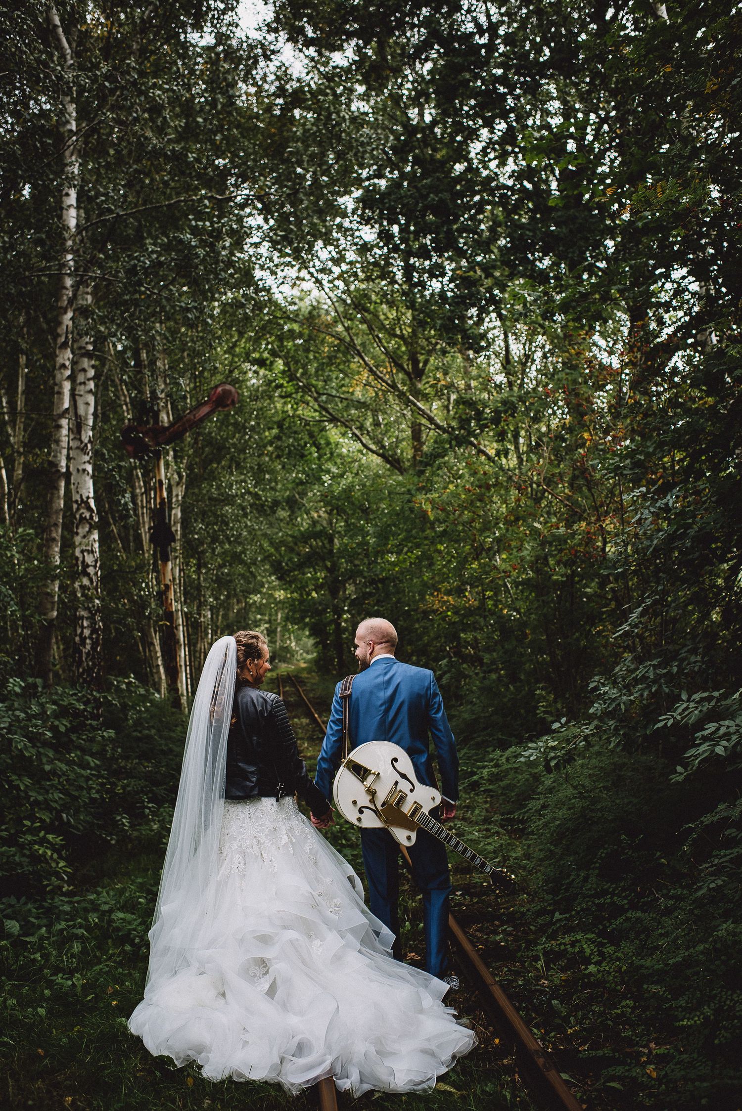 Bride in white ruffled gown and groom in blue suit walk on rails through lush green forest with guitar.
