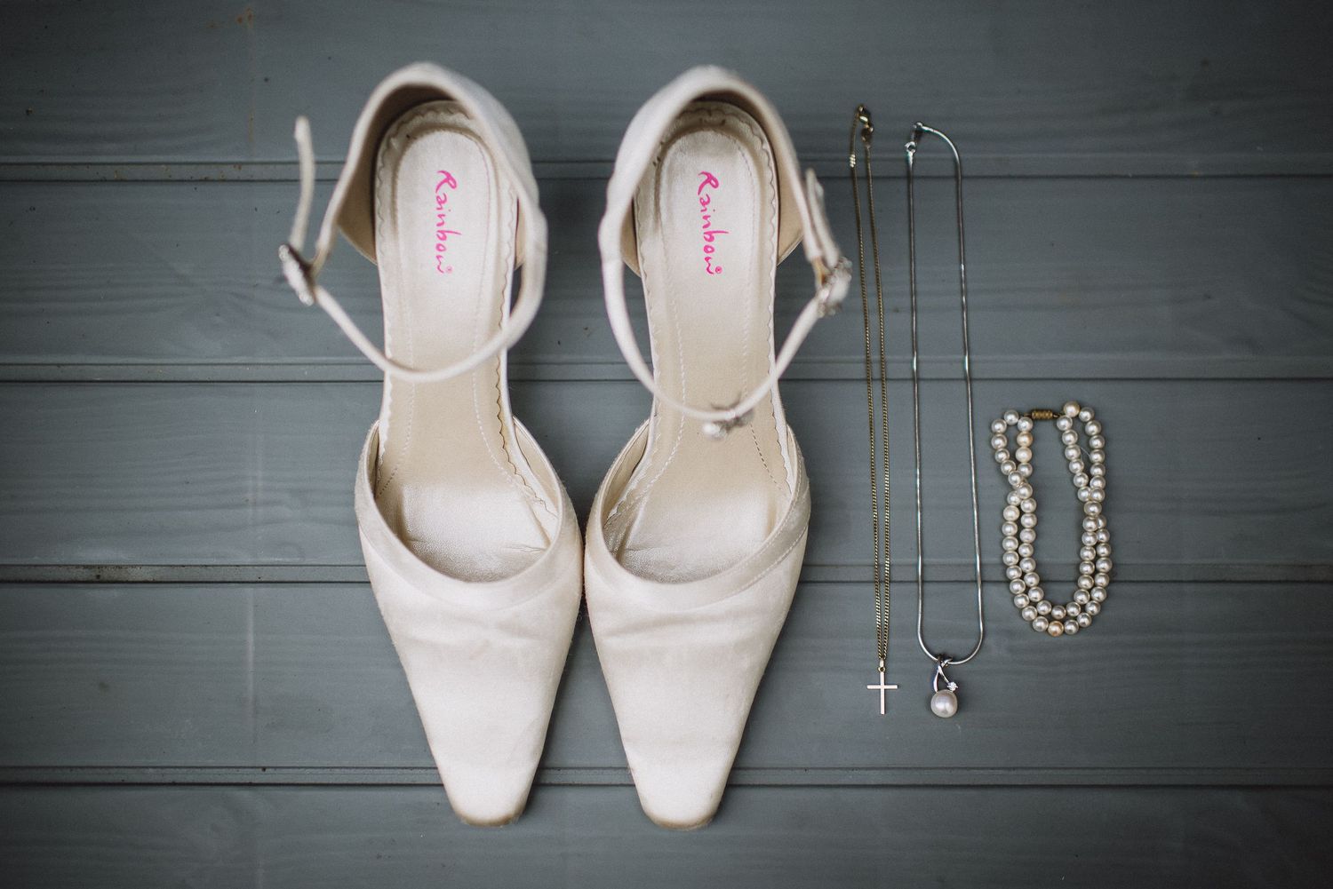 Elegant cream bridal heels displayed with pearl necklace and cross pendant on gray wood.
