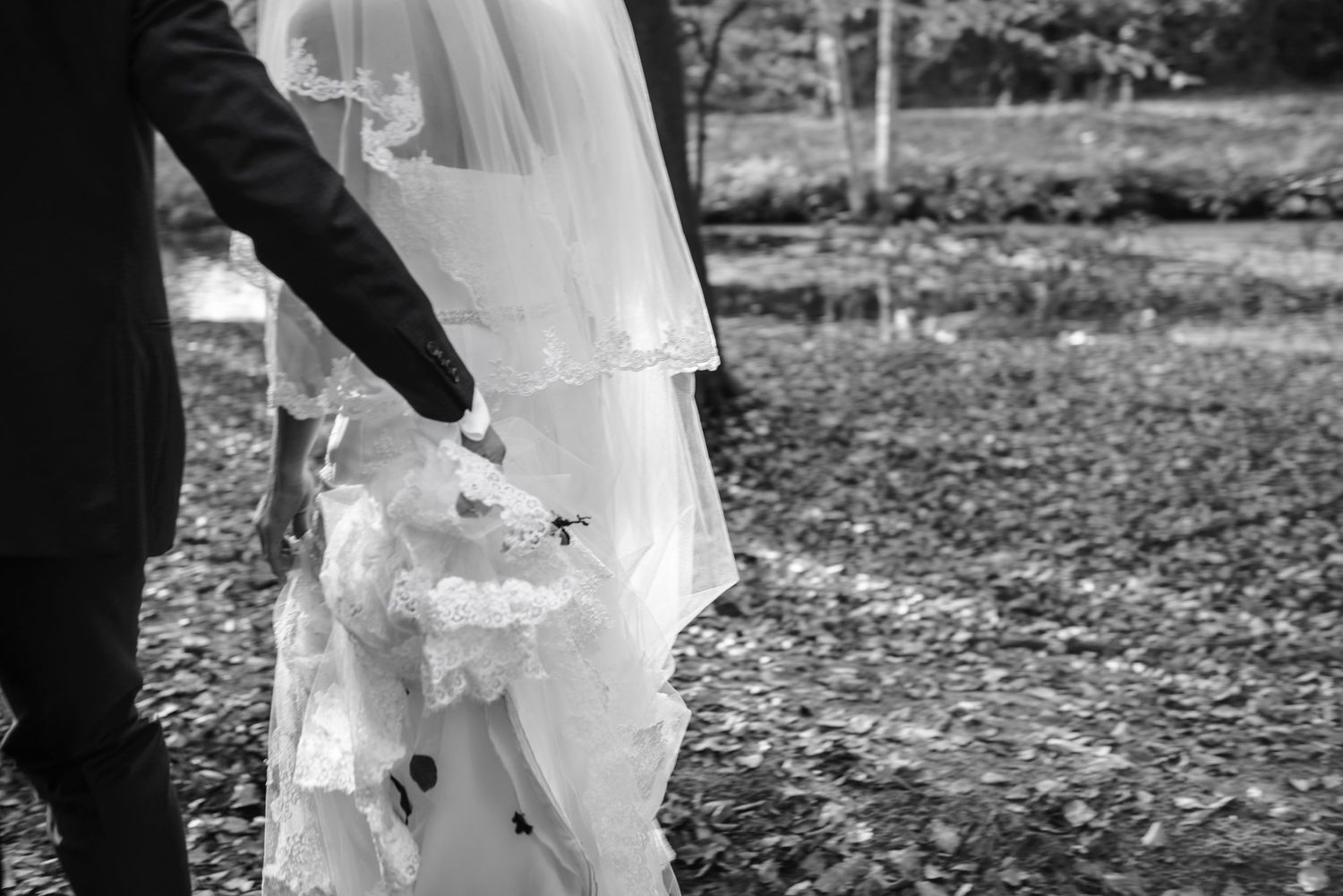 Black and white photo of a groom holding a bride's wedding dress train outdoors.