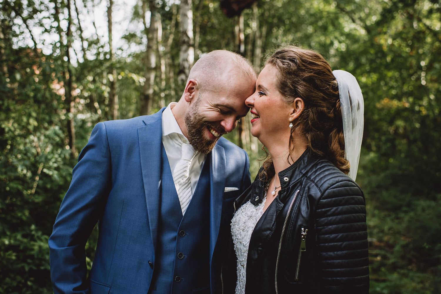 Bride in leather jacket and lace dress laughs with bald groom in blue suit forehead-to-forehead in green forest.