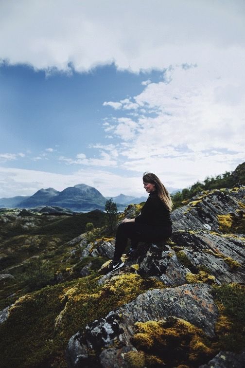 A woman in black sits on mossy rocks with a stunning mountainous Norwegian landscape behind her.