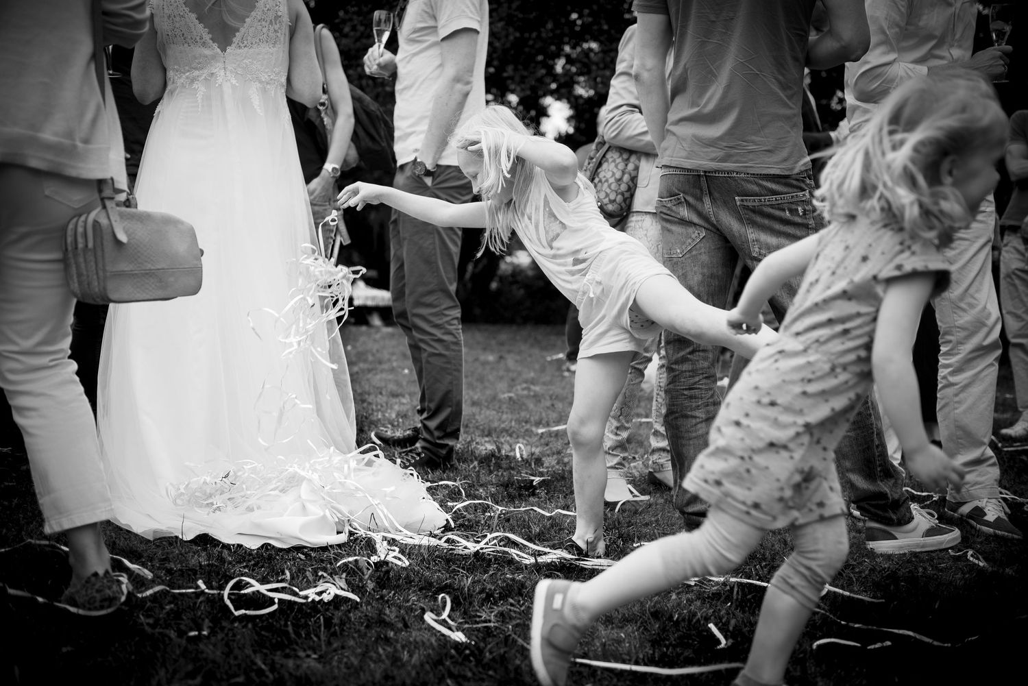 Black and white photo of children playing with streamers around bride's dress at outdoor wedding celebration.