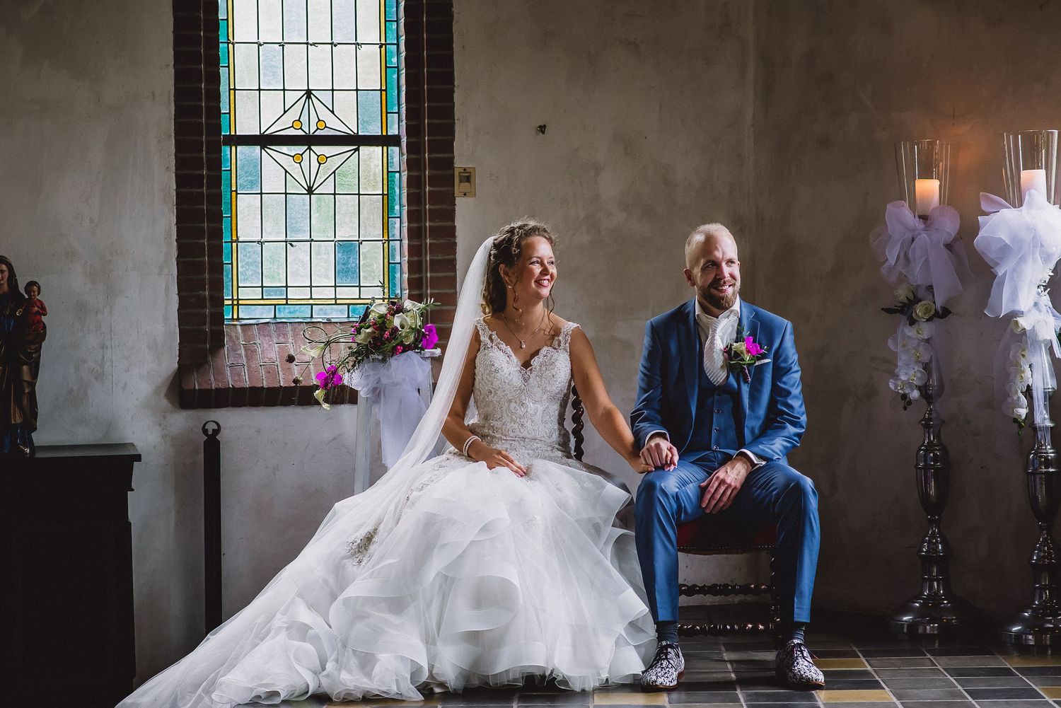 Bride and groom sit together smiling near candlelit stained glass windows in a charming church.