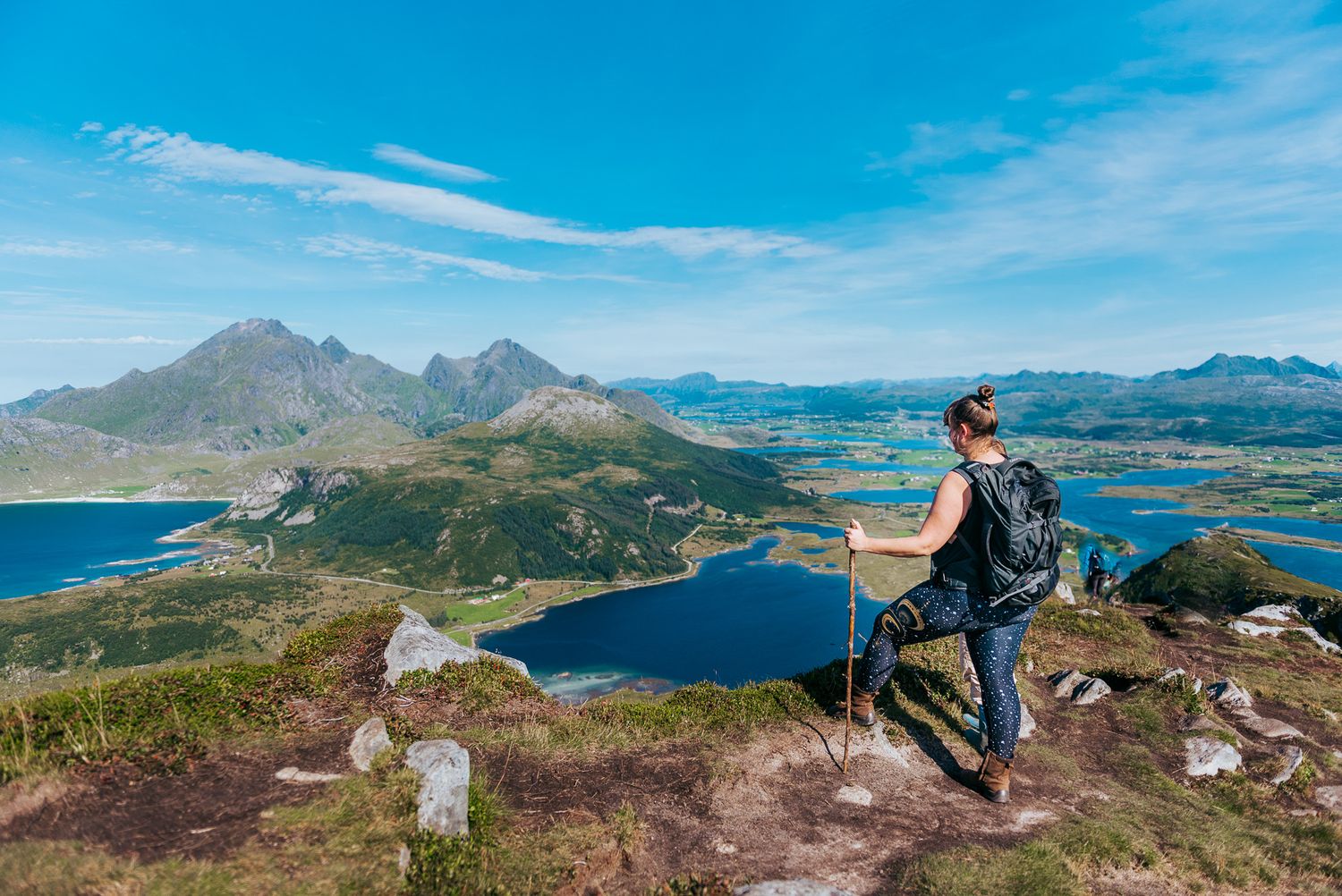 Female hiker with trekking poles standing on mountain summit overlooking Lofoten Islands archipelago, Norway.