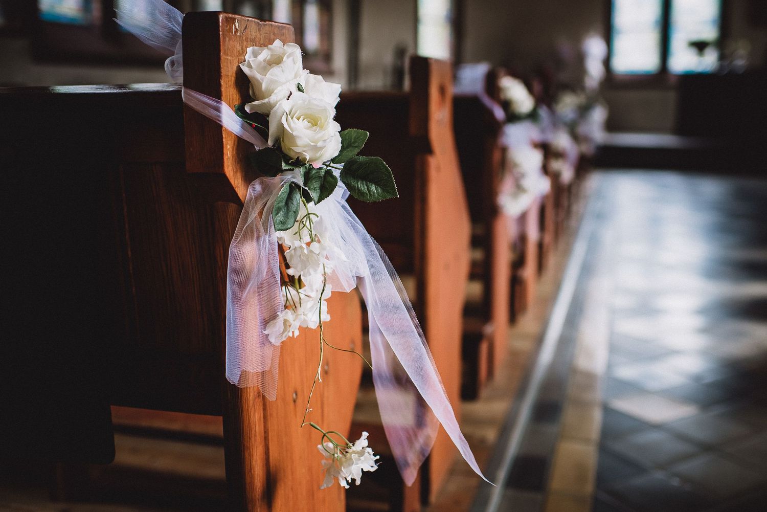 Elegant white roses and ribbon decorating wooden church pews for a romantic wedding ceremony.