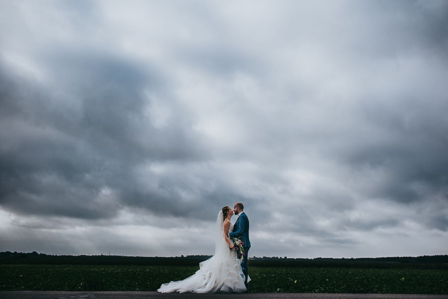 Bride and groom embrace under dramatic stormy clouds on open countryside landscape in romantic wedding portrait.