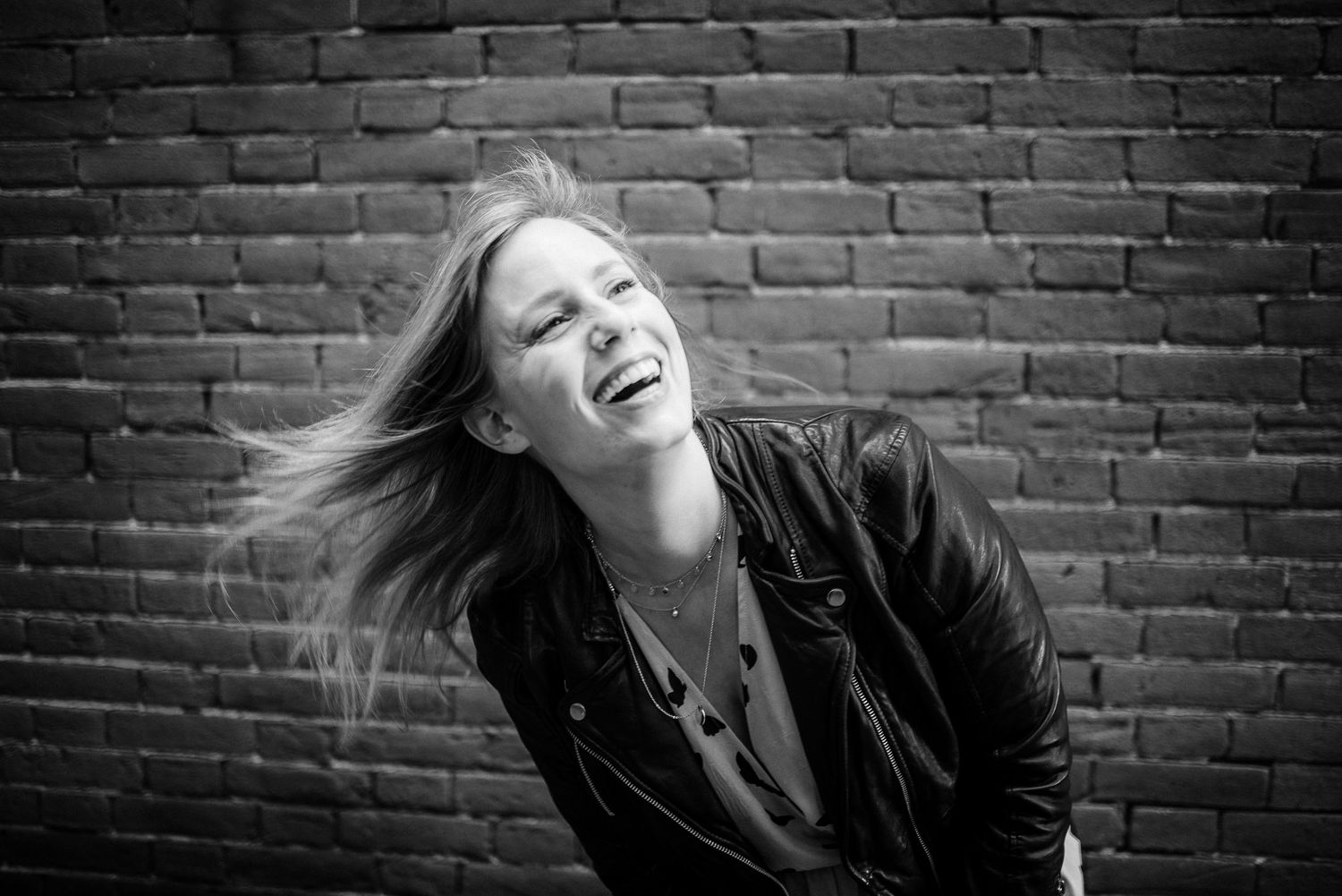 A joyful woman laughs with hair blowing in the wind against a brick wall in black and white.