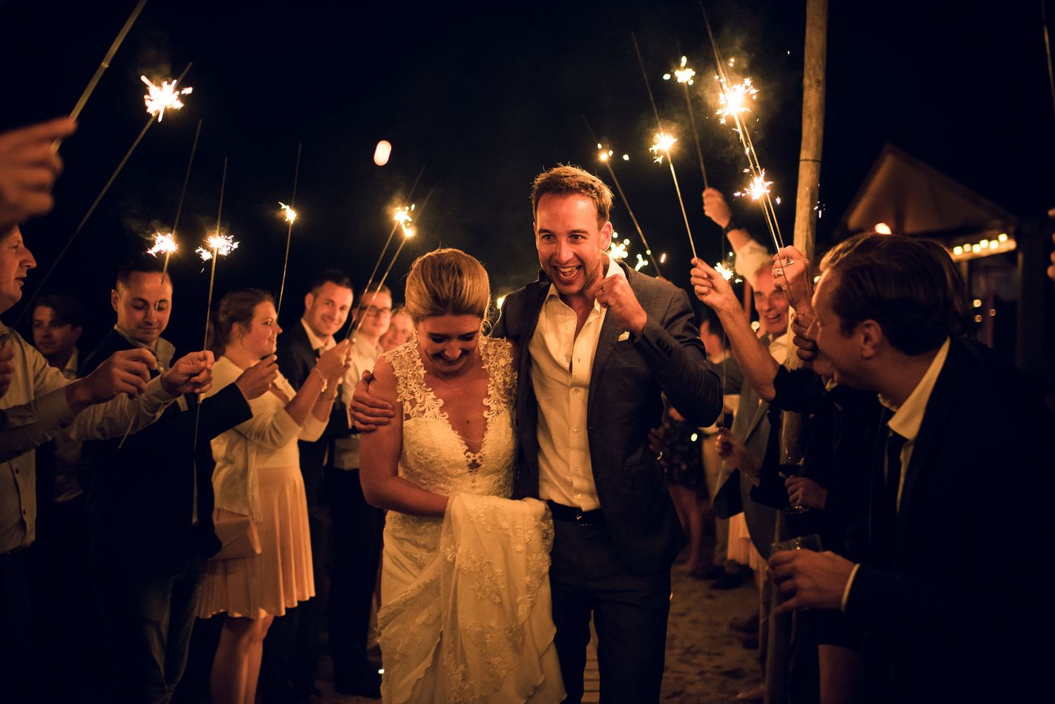 Bride and groom walking through a sparkler tunnel surrounded by cheering guests at night wedding.