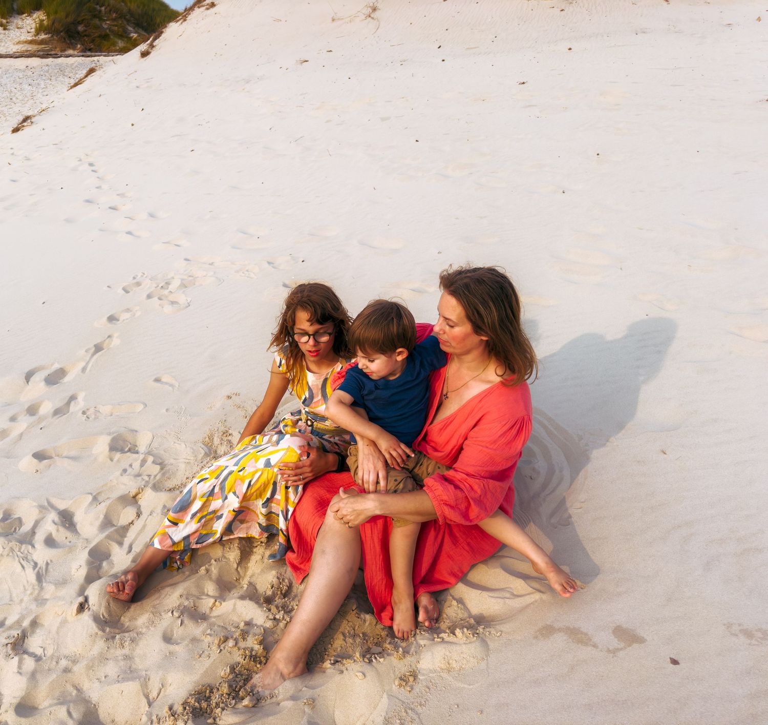 Mother sitting on white sand beach with two children in warm sunset light.