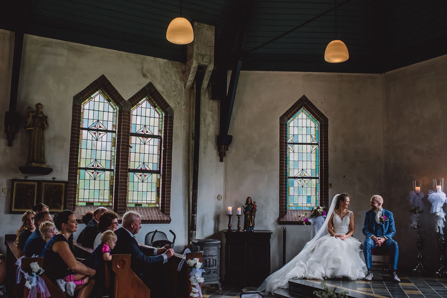 Bride in elegant gown sits near stained glass windows inside a rustic church during wedding ceremony.
