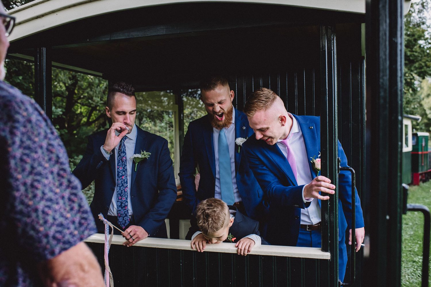 Groomsmen in navy suits laugh and socialize inside a vintage wooden trolley at a wedding. Door Kim van de Broek Fotografie