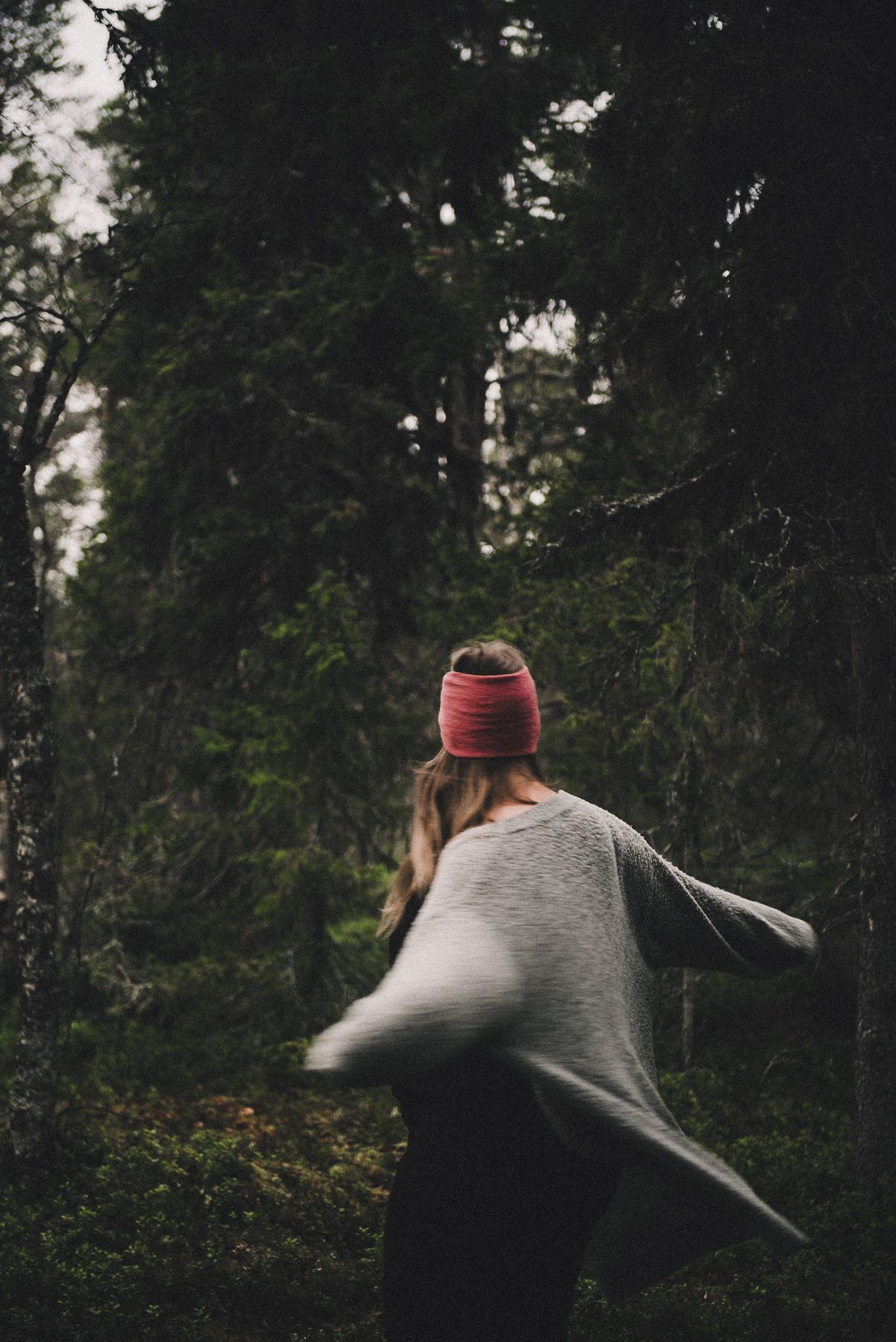 Woman in gray sweater and red beanie walking through a dark forest.