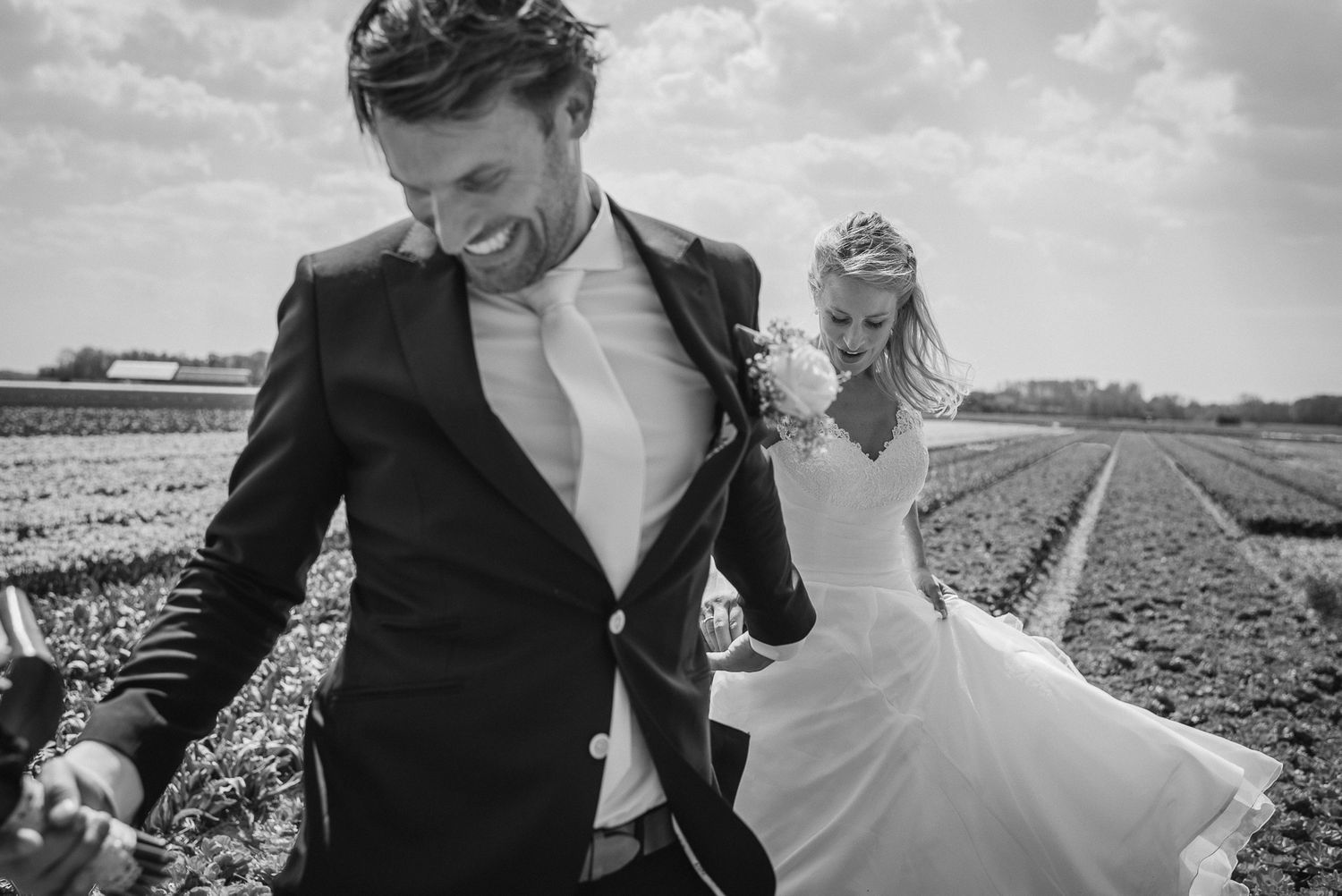 Joyful bride and groom run through a field in black and white wedding photo, bride's dress flowing.