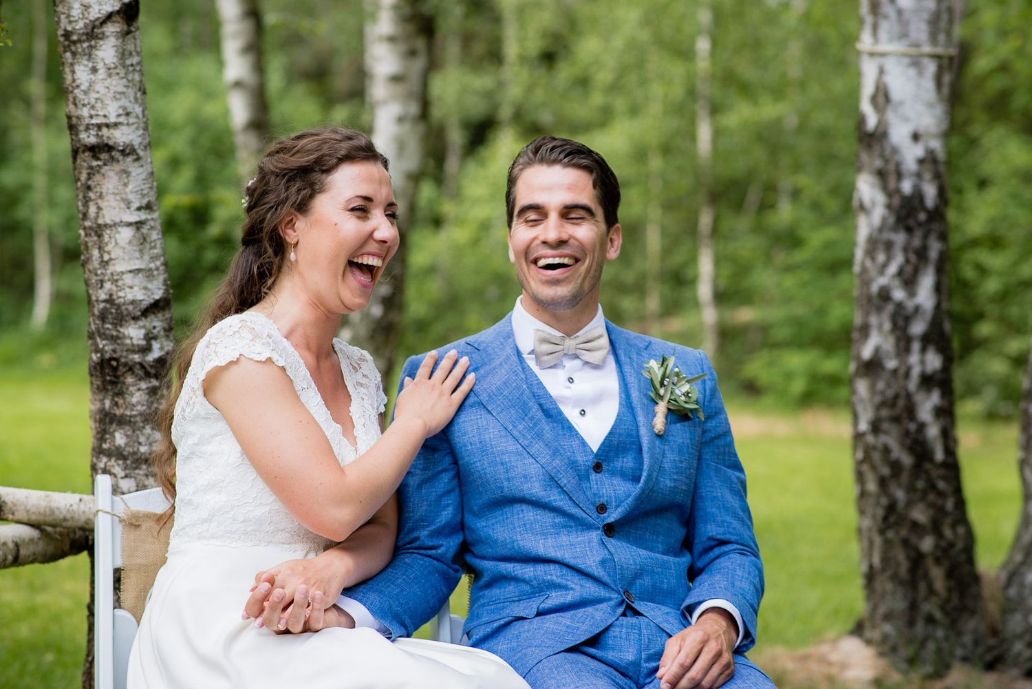 Laughing bride and groom in blue suit sitting together outdoors among birch trees on their wedding day. Door Kim van de Broek Fotografie