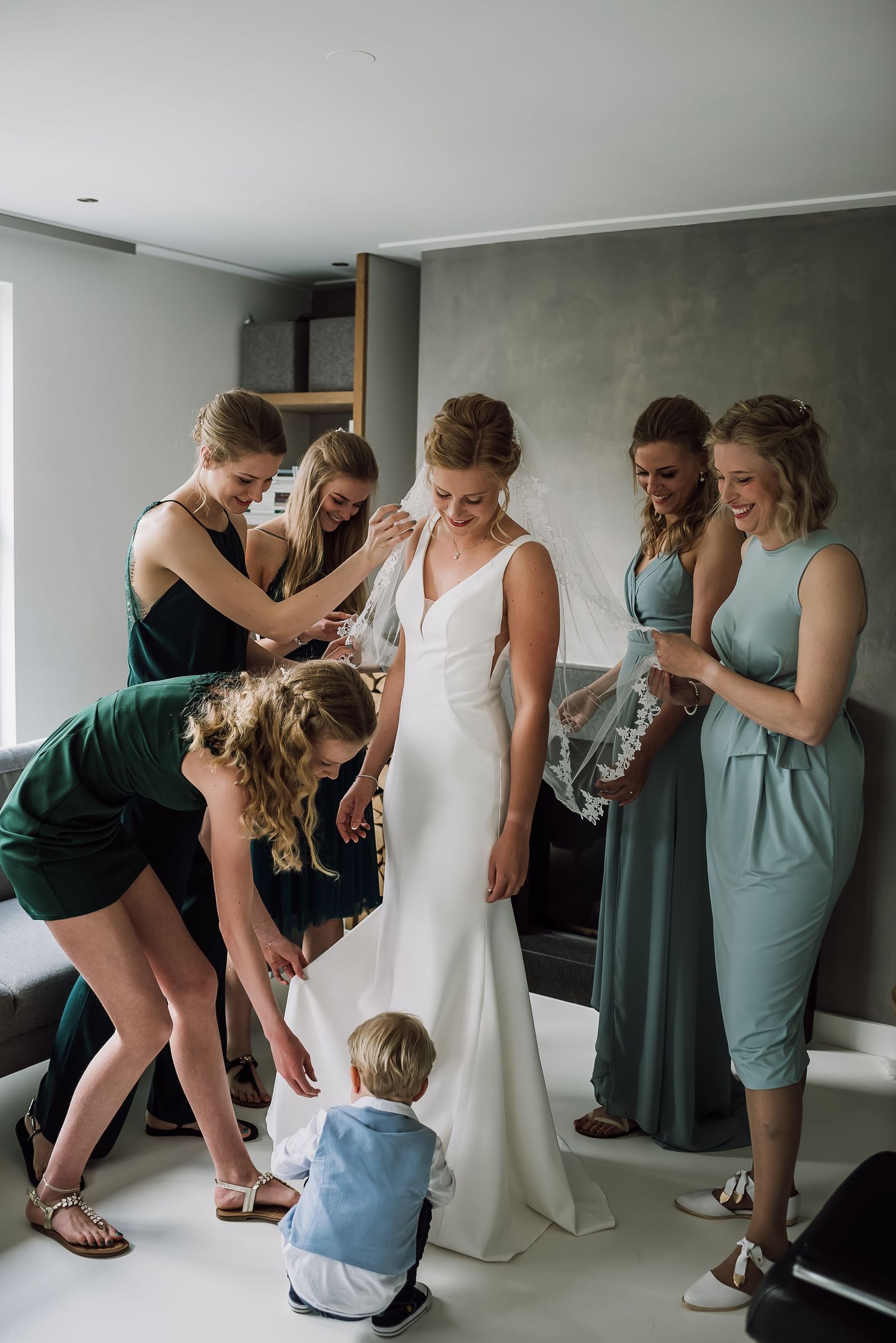 Bridesmaids in green dresses helping bride prepare while small child sits on floor nearby.