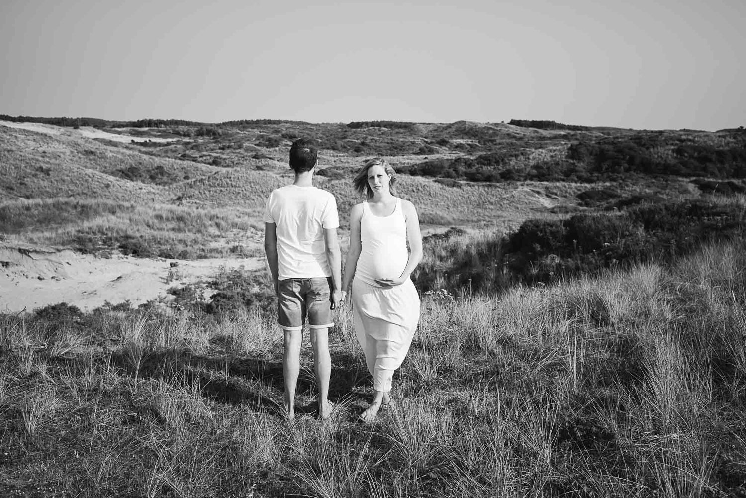 Black and white photo of a pregnant couple walking through coastal sand dunes holding hands.