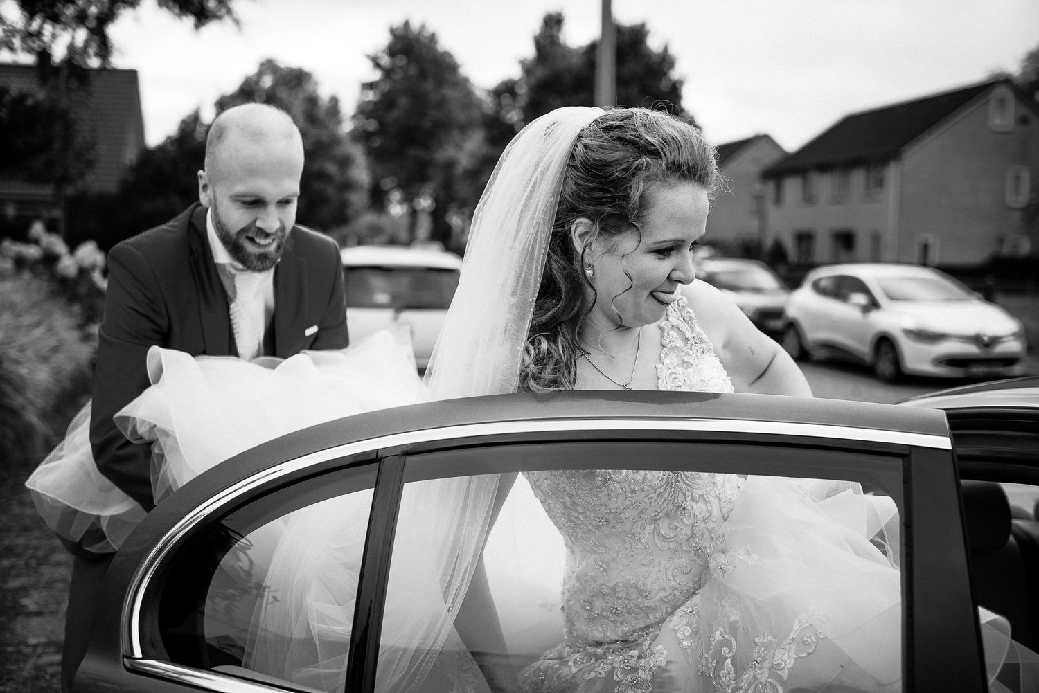 Bride in white gown and veil getting into a car as groom holds her dress in black and white wedding photo. Door Kim van de Broek Fotografie