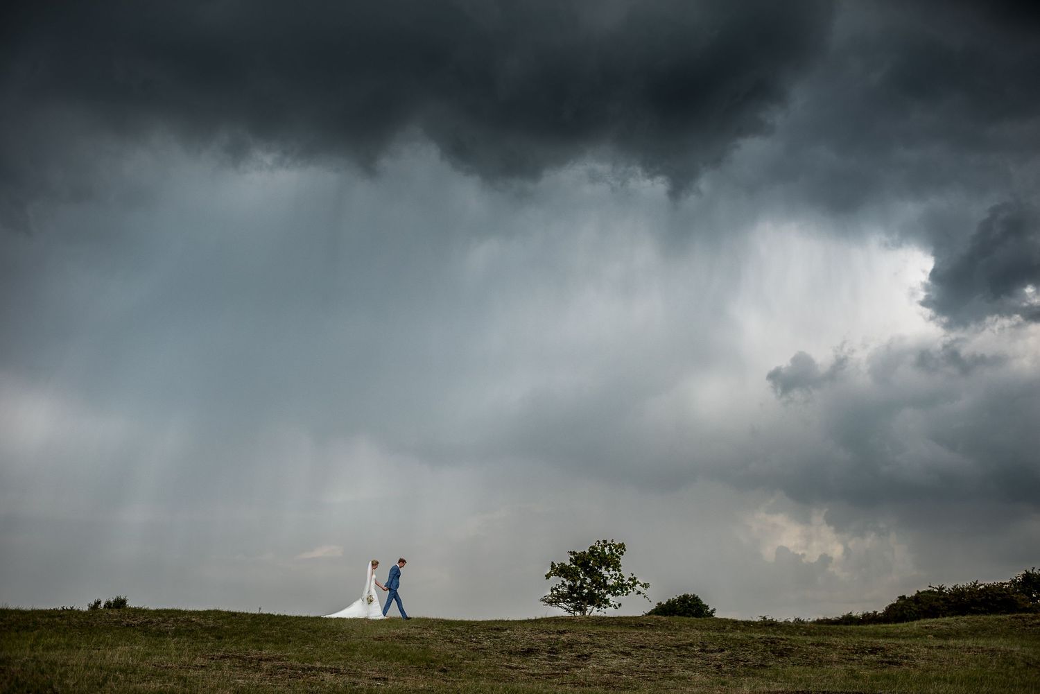 Bride and groom walk across green hillside under dramatic stormy dark clouds on their wedding day.