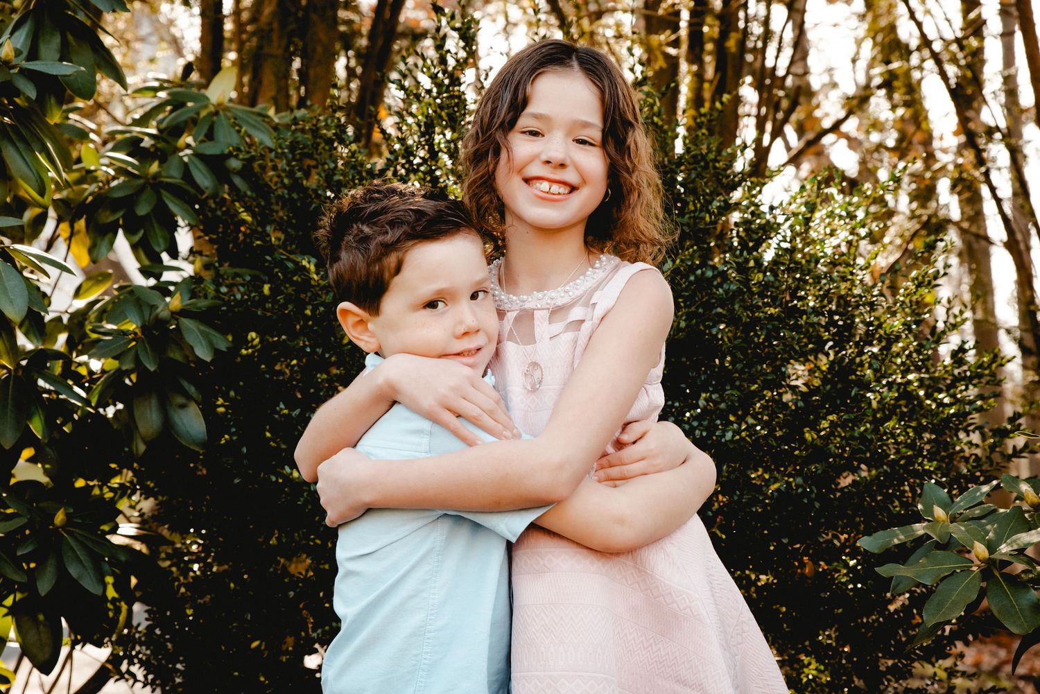 A young boy and girl hug while smiling in front of a dark green bush while the sun sets in their backyard in Ardsley, NY.