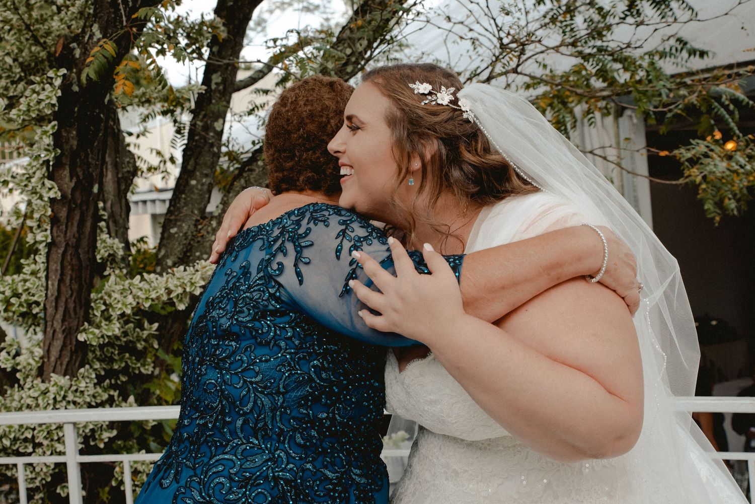 Smiling bride embraces her mother after her wedding ceremony. A close up of both women shows off the details of their beautiful dresses.