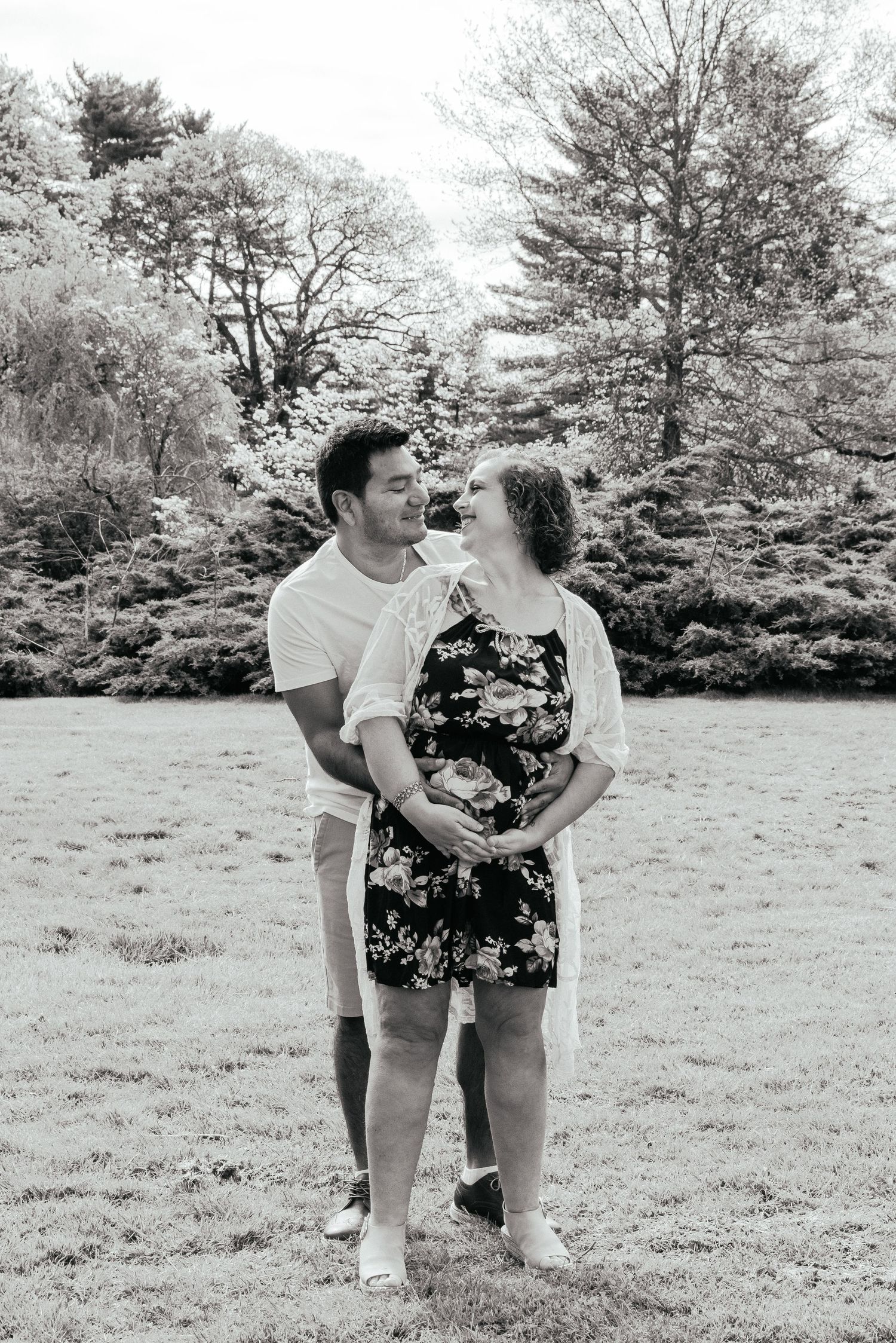 A black and white image of a man hugging his wife from behind. The couple is expecting their first child. They look at each other smiling.