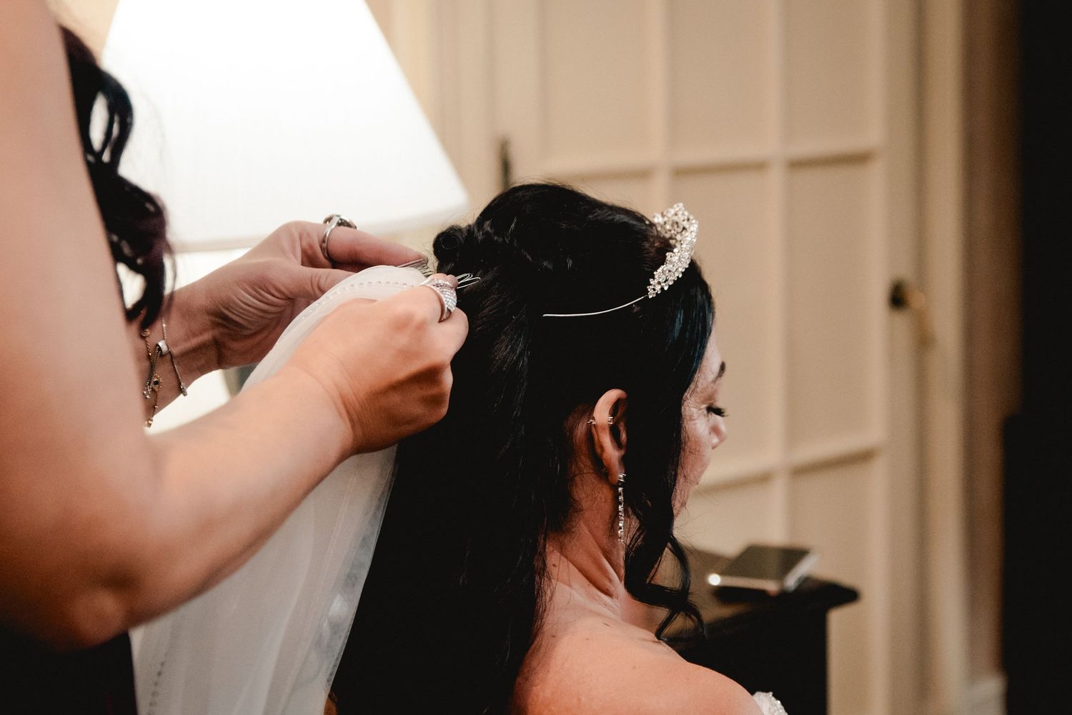 At Traditions Hotel & Spa Johnson City in Upstate NY, a bridesmaid helps the bride attach her veil to a tiara while getting ready.