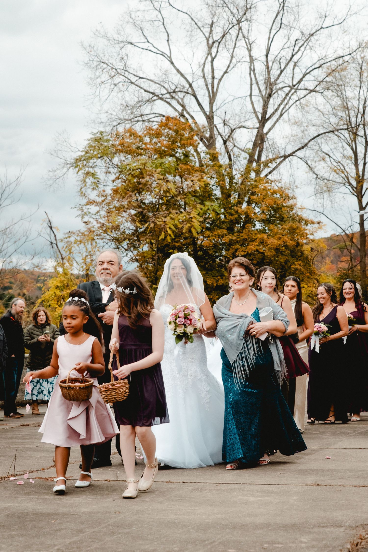 The bride is being walked to into the venue arm in arm with her parents. She is surrounded by smiling friends and family on a fall day in NY