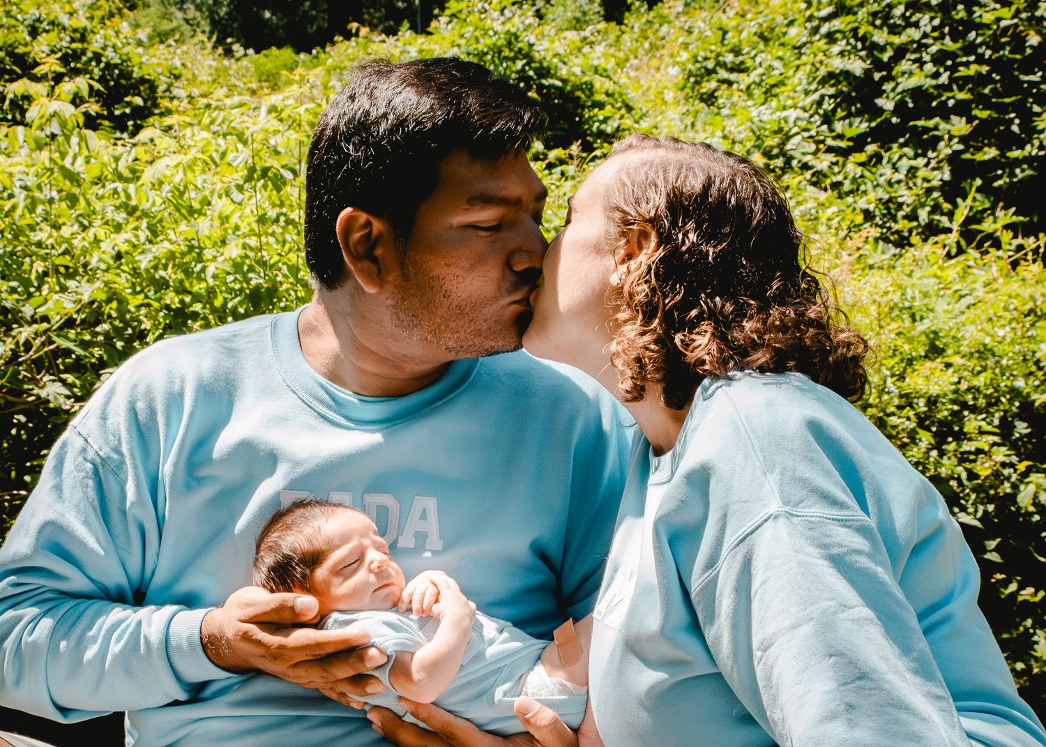 Mom & dad share a kiss while holding their infant son on a beautiful sunny day in Ardsley, NY.