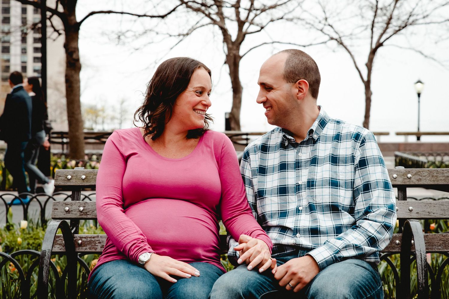 A couple expecting their first baby sits on a park bench in NYC while looking into each others eyes and laughing. She wears a pink shirt.
