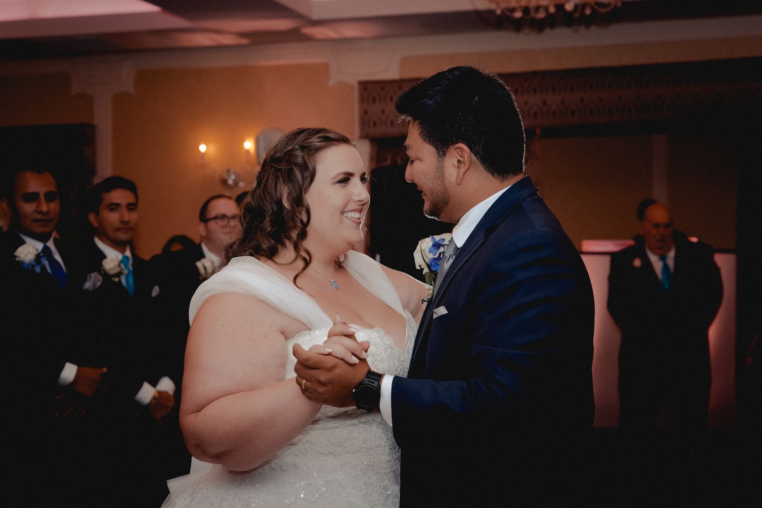 Bride and groom, both smiling, share their first dance surrounded by friends and family. The room is softly lit, resembling candlelight.